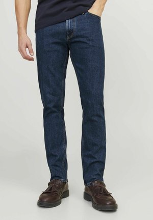 Homme portant un jean slim bleu foncé et des mocassins en cuir marron foncé à pampilles, debout avec une main dans la poche.