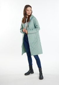 Cappotto lungo trapuntato di colore verde chiaro, dotato di cappuccio, chiusure a scatto e taglio dritto. Indossato sopra un top grigio e jeans blu con stivali neri.