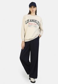 Sudadera beige con el texto "LOS ÁNGELES" y detalles rojos, combinada con pantalones sueltos azul marino y zapatos grises, rematada con una gorra azul marino.