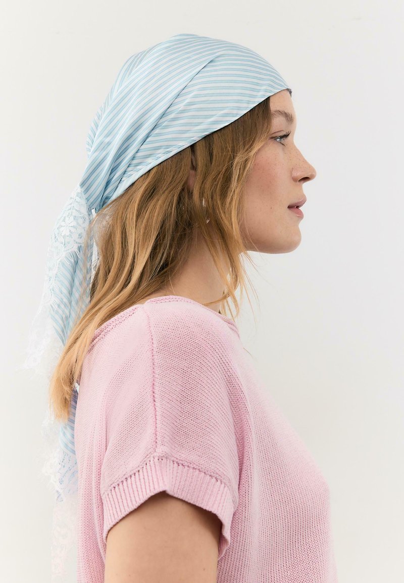 Femme de profil portant un foulard à rayures bleu clair avec de la dentelle et un haut tricoté rose à manches courtes, sur un fond uni.