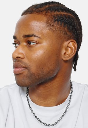 Profil d'une personne avec des cheveux tressés, portant un t-shirt blanc et un collier en chaîne en argent épais. Peau lisse, arrière-plan neutre.