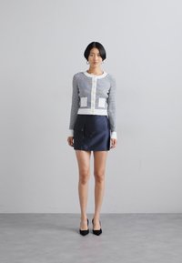 Strikket cardigan med marineblå og hvite striper, hvite detaljer og to frontlommer, kombinert med et marineblått miniskjørt. Svarte høye hæler.