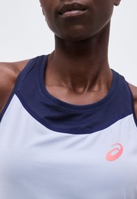 Κοντινό πλάνο ατόμου που φορά αθλητικό μπλουζάκι χωρίς μανίκια σε σκούρο μπλε και άσπρο με ροζ λογότυπο Asics στο στήθος, μπροστά από απλό φόντο.