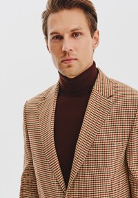 Brązowa i beżowa marynarka w wzór houndstooth z klapami, noszona na ciemno-brązowym golfie, z teksturowanej tkaniny, dopasowany krój.