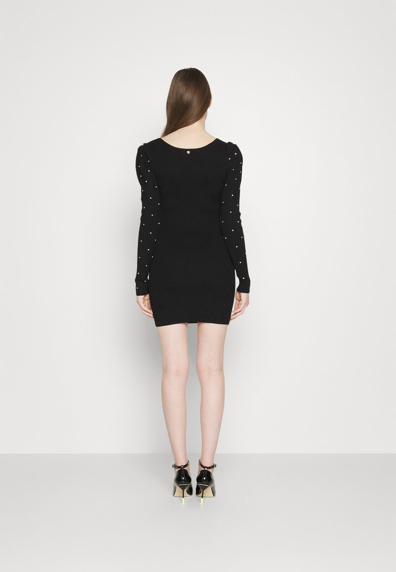 Schwarzes Minikleid mit langen, schmalen Ärmeln, die mit perlenähnlichen Akzenten verziert sind. Glatte Stricktextur und runder Ausschnitt. Das Model trägt schwarze Absätze mit gelben Schuhen.