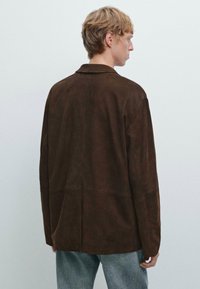 Mokka suède jas met een relaxed fit, notch kraag en rugpendetail, gecombineerd met lichtblauwe jeans. De textuur is glad en mat.