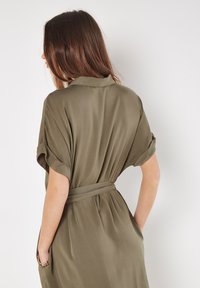 Olivegrünes, kurzärmliges Kleid aus weichem Stoff, mit einem Bindegürtel in der Taille und seitlichen Taschen. Angenehme Textur und minimalistisches Design.