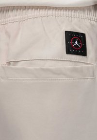 Beige byxor med elastisk midjeband, med en bakficka och en svart etikett med en röd cirkel och vit logotyp på övre vänstra området.