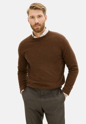 Maglione marrone realizzato in materiali morbidi, colletto rotondo, polsini a costine, abbinato a pantaloni grigi a fantasia. Modella in piedi con le mani in tasca.