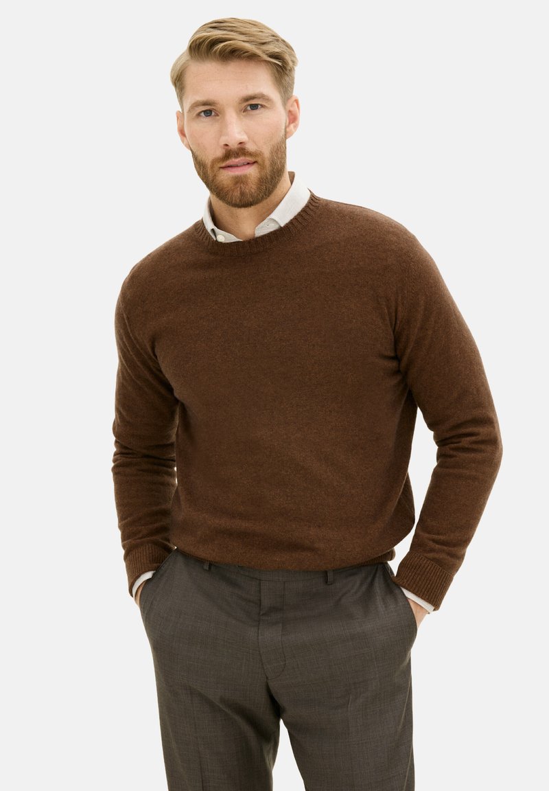 Maglione marrone realizzato in materiali morbidi, colletto rotondo, polsini a costine, abbinato a pantaloni grigi a fantasia. Modella in piedi con le mani in tasca.