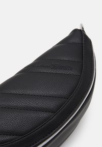 Černý kožený pouzdro s zakřiveným tvarem, které má prošívanou texturu a stříbrný zip. Na povrchu je viditelný embossovaný detail loga.