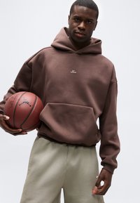 Άνδρας που φοράει καφέ φούτερ με κουκούλα και μπεζ παντελόνι κρατώντας μια μπάλα μπάσκετ μπροστά σε απλό φωτεινό φόντο.