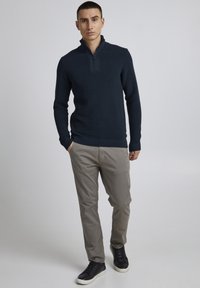 Tumesinine pikkade varrukatega pullover poolsiidiga kraega, ribakujulise tekstuuriga, koos heleda khaki pükste ja mustade valge põhjaga tennisetossudega.