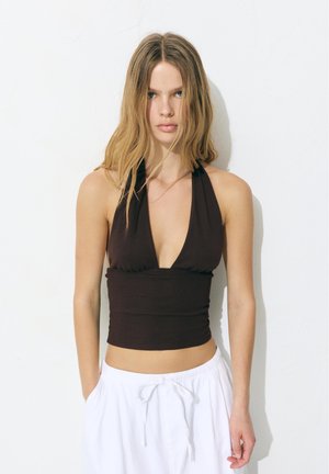 Młoda kobieta z długimi blond włosami, ubrana w czarny top z wiązanym tyłem i białe spodnie ze sznurkiem, stoi przy białej ścianie.