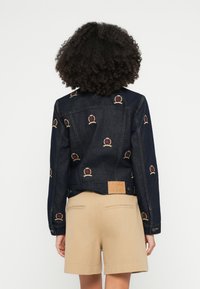 Tmavomodrá rifľová bunda s farebnou výšivkou a koženou značkou v hnedej farbe. Štýlovo skombinovaná s béžovými šortkami, viditeľnými kovovými prvkami a detailmi šitia.