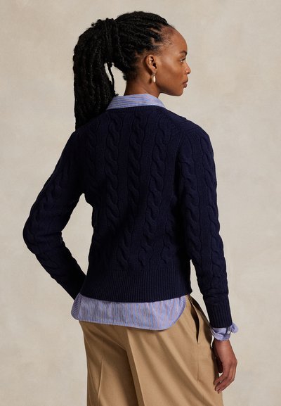 Mujer con el pelo trenzado recogido en una coleta lleva un suéter azul marino de punto con ochos sobre una camisa de rayas y pantalones beige, de pie frente a un fondo liso.