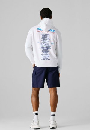 Hombre con sudadera con capucha blanca con las palabras "World Tour Red Bull Racing" y una lista de ciudades globales impresas en la espalda, pantalones cortos azul marino, calcetas blancas y zapatillas deportivas.