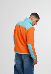 Giacca in pile con corpo arancione e spalle turchesi, dotata di un colletto alto. Sul retro appare un piccolo logo e un tessuto testurizzato.