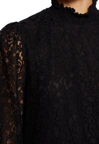 Top in pizzo nero caratterizzato da un collare alto con bordo arricciato, texture trasparente e motivo floreale, che mette in evidenza i dettagli intricati sulle maniche.
