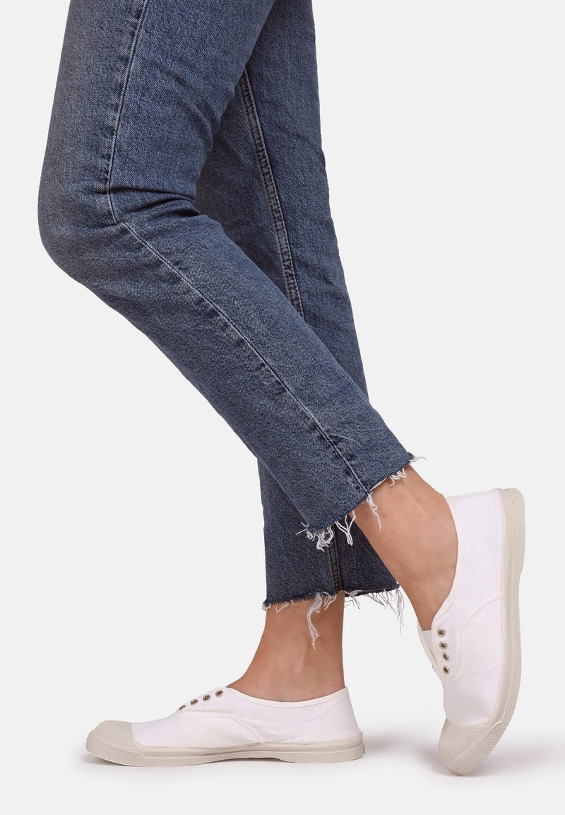 Baskets à enfiler en toile blanche avec des semelles en caoutchouc beige et des œillets en métal tricolore, associées à un jean en denim foncé avec un ourlet effiloché.