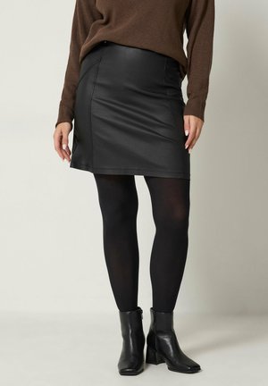 Frau trägt einen schwarzen Lederrock, schwarze Strumpfhosen, schwarze Stiefeletten und einen braunen Langarmpullover und steht vor einem einfarbigen Hintergrund.