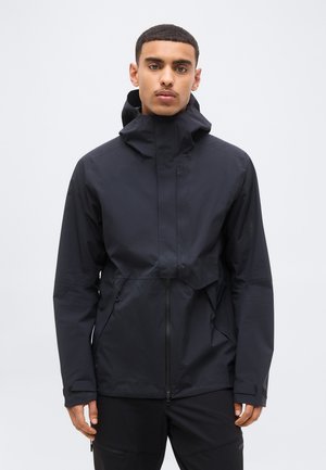 Young man wearing a black hooded jacket with front zipper and zippered side pockets, standing against a plain light background.