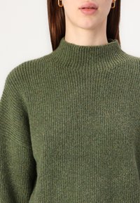 Olijfgroen geribbeld gebreid sweater met een hoge kraag, met een dikke textuur en een effen kleur die de zachtheid en warmte benadrukt.