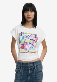 Weißes Baumwoll-T-Shirt mit kurzen Ärmeln, das mit einem bunten Grafikdesign aus Herzen und Mustern versehen ist, kombiniert mit hoch taillierten Jeansshorts.