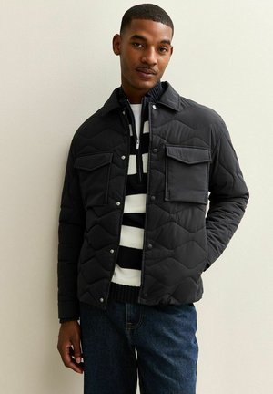 Black quilted jacket with snap buttons, two chest pockets, and a textured finish, worn over a striped black and white top and dark jeans.