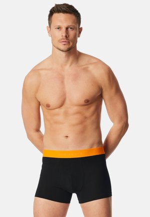 Muskuløs mand stående med armene bag ryggen, iført sorte boxershorts med orange talje mod en ensfarvet baggrund.