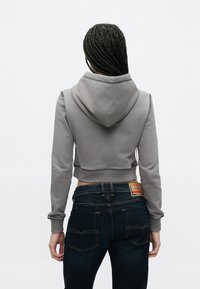 Šedá skrátená mikina s kapucňou, vybavená rebrovanými manžetami a surovým lemom, v kombinácii s tmavými džínami s viditeľnými detailmi prešívania.