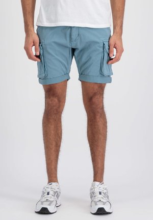 Homme portant un short cargo bleu clair et des baskets blanches, se tenant devant un fond blanc uni.