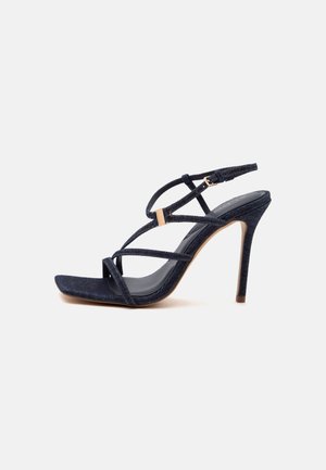 Dunkelblaue Sandalette mit hohem Absatz, dünnen gekreuzten Riemen und einer kleinen goldenen Schnalle auf weißem Hintergrund.