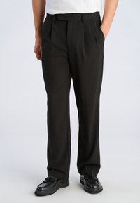 Person iført sorte bukser med lige ben og sidelommer samt sorte loafers med kvaster, stående mod en enkel, lys baggrund.