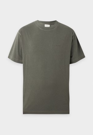 Mann mit rasiertem Kopf, der ein dunkelgraues kurzärmeliges T-Shirt, beige weite Hose und schwarze Schuhe trägt, steht mit einer Hand in der Tasche.