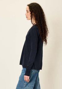 Moteris su ilgais garbanotais plaukais, vilkinti tamsiai mėlyną ilgomis rankovėmis marškinius ir mėlynus džinsus, stovinti profilio požiūriu prieš šviesią foną.