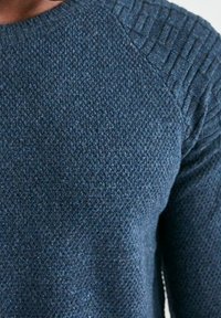 Marinblå stickad tröja med texturerad kropp och ribbad axeldetalj. Tyget verkar tjockt och varmt, lämpligt för svalt väder.