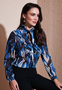 Sheer blouse featuring blue, black, and white wave patterns, ruffled detailing, a high collar, and a front tie. Black fitted pants.
