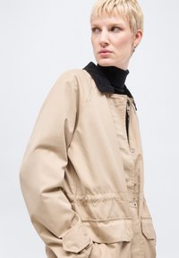 Ξανθό άτομο με μπεζ μπουφάν με μαύρο γιακά και μαύρο ζιβάγκο, που κοιτάζει στο πλάι με σοβαρή έκφραση πάνω σε λευκό φόντο.