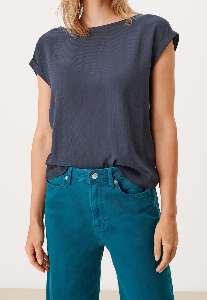 Femme portant un chemisier à manches courtes bleu marine foncé rentré dans un pantalon taille haute couleur sarcelle avec poches avant et fermeture par boutons.