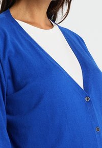 Niebieski kardigan wykonany z miękkiego materiału; otwarty przód z głębokim dekoltem w serek, z dwoma małymi guzikami i gładką fakturą.