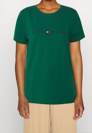 Person wearing a green Tommy Hilfiger t-shirt with logo and "EST. 1985" text, paired with beige ribbed pants, against a neutral background.