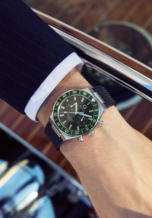 Mandens håndled i en mørk pinstripe-dragt iført et Timex kronografur med sort urskive, grøn bezel og sort tekstureret rem.