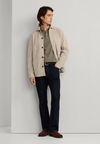 Muškarac u bež zakopčanom saku, maslinasto zelenoj polo majici, tamnim trapericama i smeđim cipelama stoji naslonjen na svijetlosivu zidnu površinu, gledajući bočno.