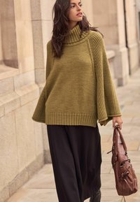 Oversized olive green knit sweater with a high ribbed collar and wide sleeves, paired with a long black skirt and a brown handbag.
