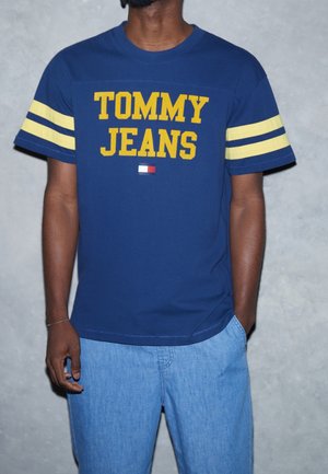 Mies pukeutunut laivaston siniseen "TOMMY JEANS" -t-paitaan, jossa on keltaiset raidat hihoissa, ja vaaleansiniset farkkushortsit, seisoo harmaata seinää vasten.