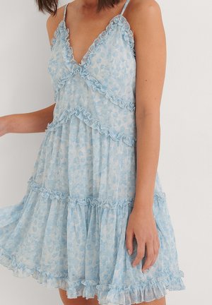 Femme portant une robe d'été fleurie bleu clair avec un bord volanté et des bretelles fines, debout contre un mur blanc.