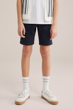 Partie inférieure d'une personne portant un short bleu marine, des chaussettes blanches avec des rayures grises, des baskets blanches à semelles en gomme, une chemise blanche et un gilet crème.