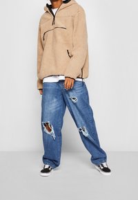 Pullover in pile beige con cappuccio e dettagli neri, abbinato a jeans oversize blu con ampie zone strappate. Sneakers nere completano il look.