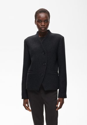 Femme portant un blazer noir cintré à trois boutons et un pantalon noir assorti, debout devant un fond blanc uni.